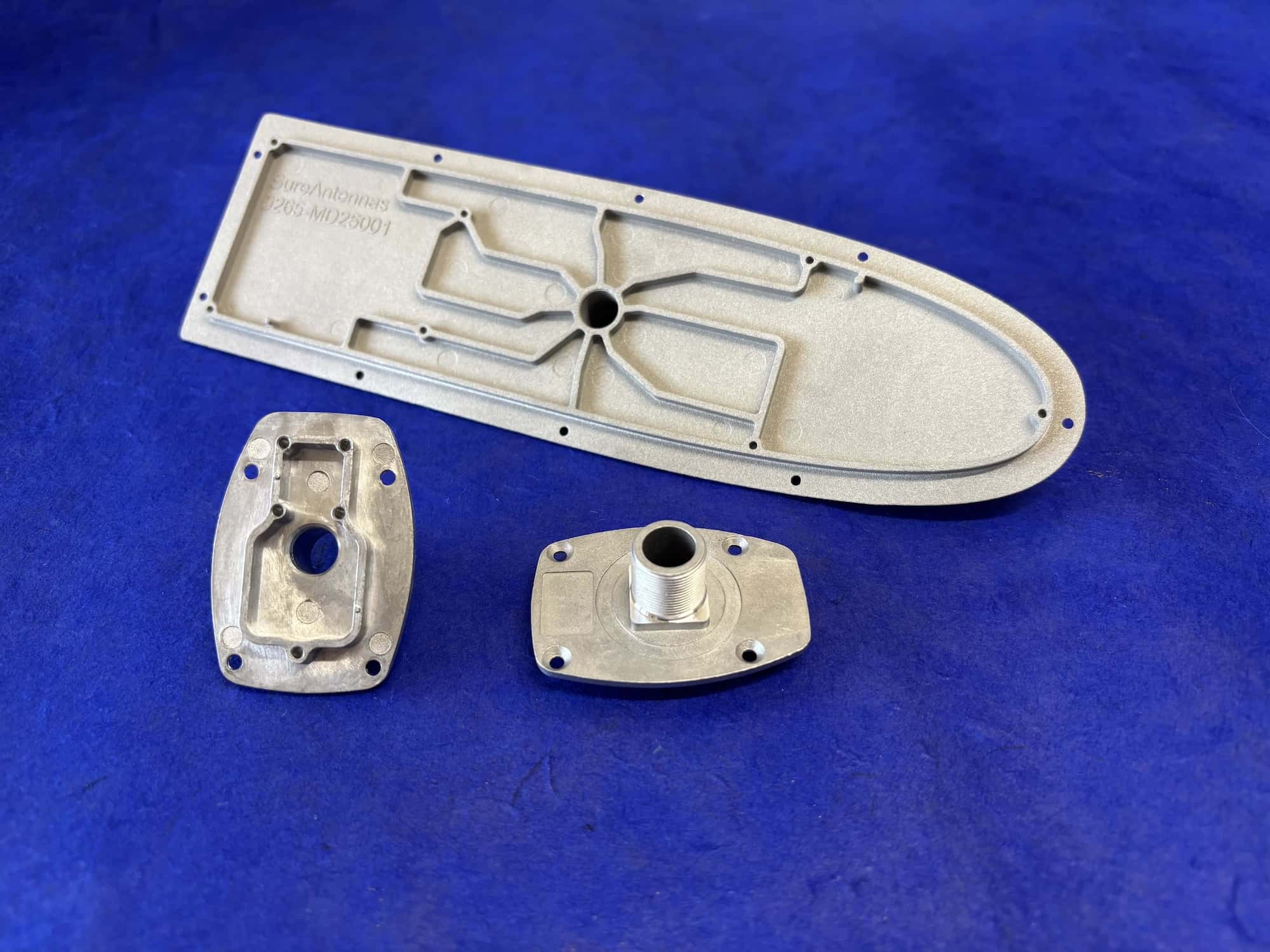 Three aluminium pressure die-cast components on a blue background, including an elongated channelled base plate and two smaller housings